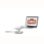 Imagem do scanner-intra-oral-trios 3shape ao lado de um laptop que exibe uma modelagem de dentes em alta qualidade e precisão.