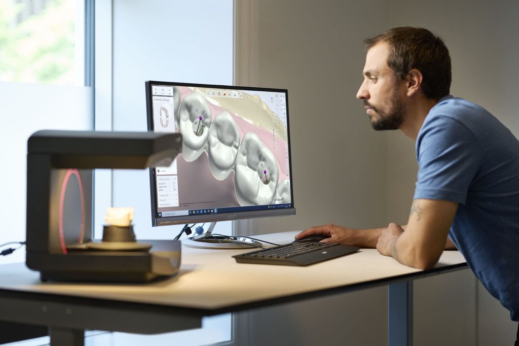 Técnico de Prótese Dentária com um scanner de bancada e um computador em sua frente, no computador uma imagem de software aberta com alguns dentes na tela e o profissional observando.