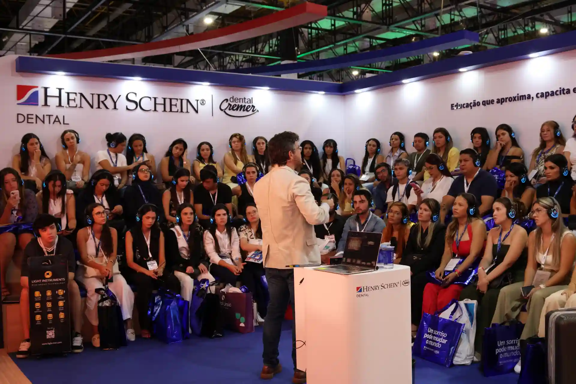Palestrante apresentando para um grande público em evento de odontologia da Henry Schein, com diversas pessoas assistindo atentamente e usando fones de ouvido.