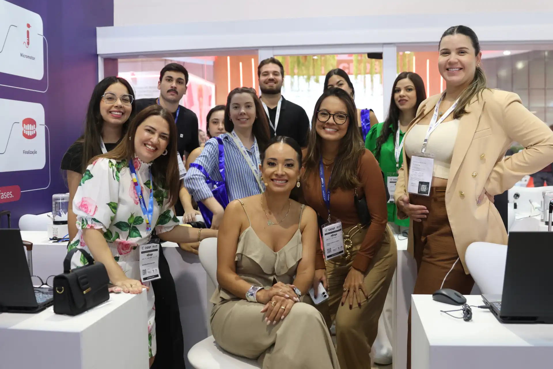 Grupo de pessoas sorrindo na feira de negócios, participando de networking e eventos corporativos, com destaque para mulher de vestido bege no centro da imagem.