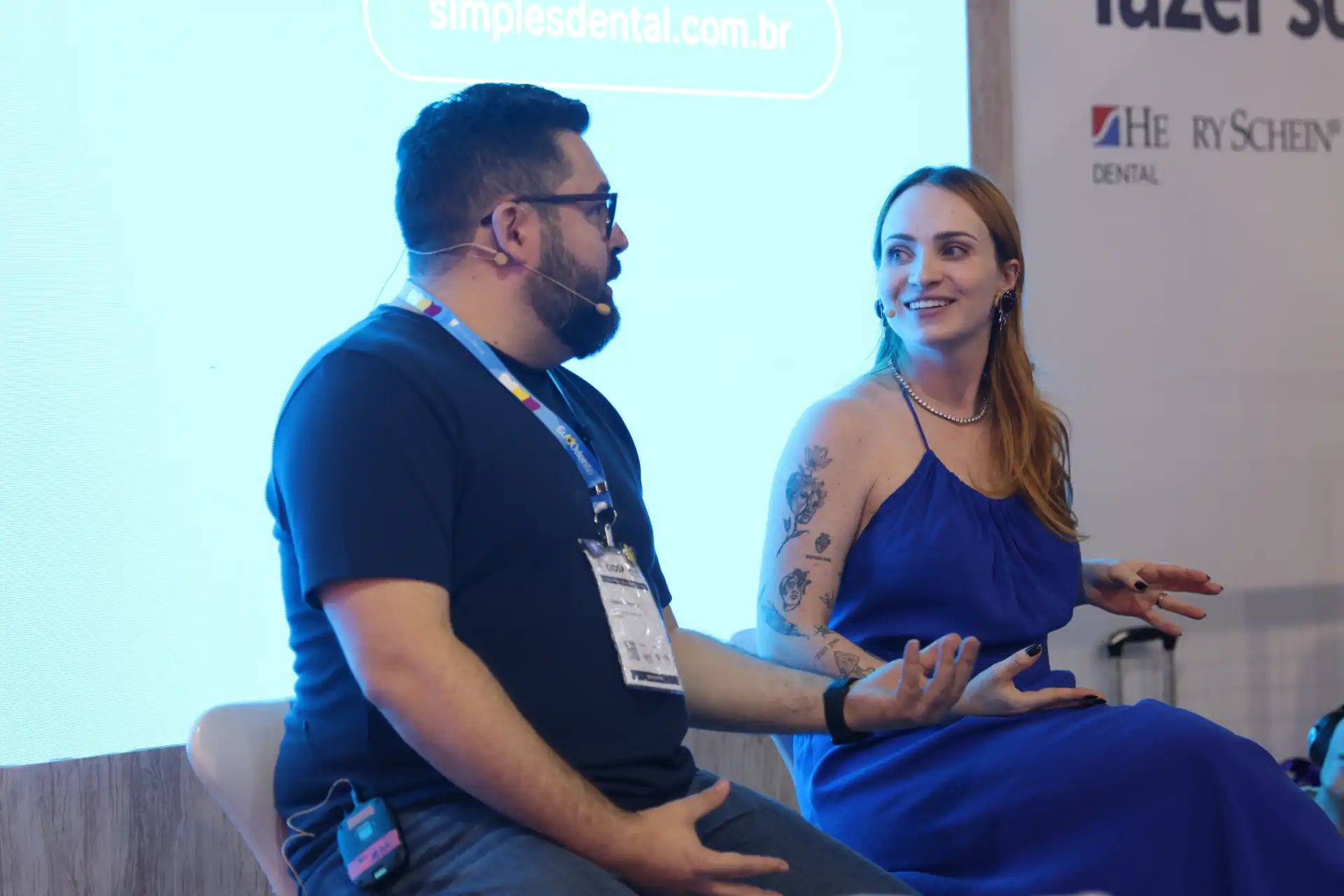 Dois profissionais conversando durante um evento, um homem de barba, óculos e camiseta escura e uma mulher de cabelo castanho, tatuagens e vestido azul, sentados em palco com tela ao fundo.