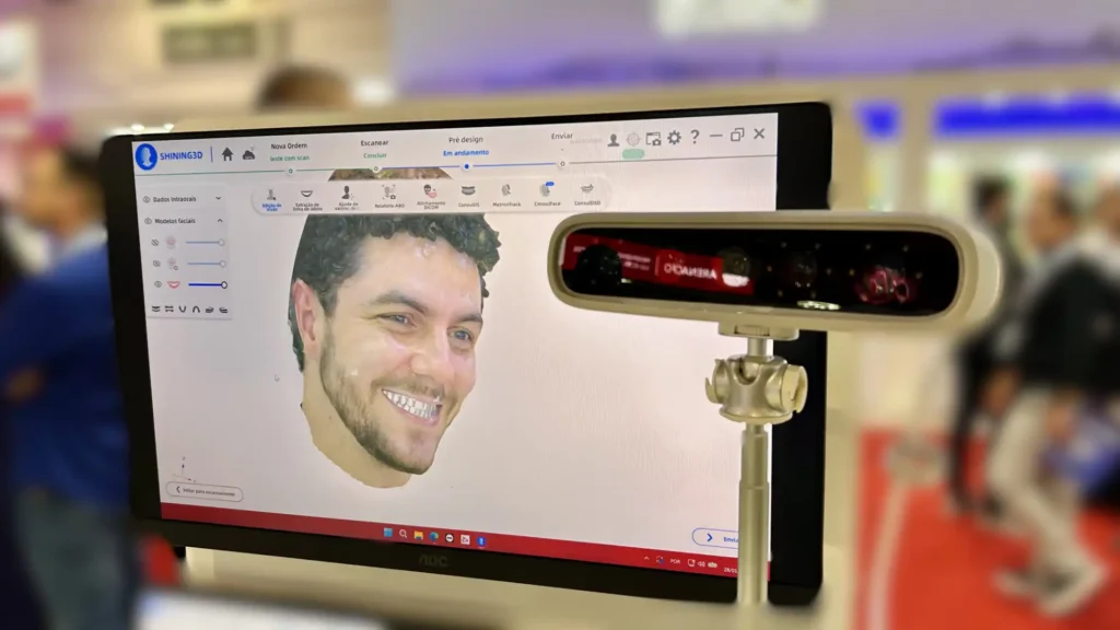 Homem sorridente em frente a um computador na feira de tecnologia, com reconhecimento facial e inova&ccedil;&atilde;o digital em destaque.