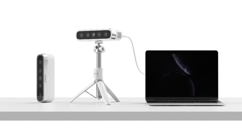 Equipamentos de escaneamento facial, incluindo uma c&acirc;mera 3D, um projector port&aacute;til, um computador port&aacute;til e uma base trip&eacute;, todos dispostos em uma mesa branca.